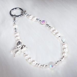 May include: A pearl beaded keychain with a silver bow charm and iridescent crystal accents. The keychain features a silver ring and small silver beads interspersed with the pearls. The background is a soft, white, fluffy surface.