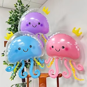 May include: Three colorful jellyfish-shaped balloons. The balloons are in shades of blue, pink, and purple, each with a smiling face and trailing tentacles. Yellow crown-shaped decorations are also present, suggesting a party or celebration theme.