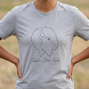 May include: A heather gray short-sleeve t-shirt featuring a black line art illustration of a bison head centered on the chest. Below the bison graphic, the text Custer State Park is printed in a simple, dark, cursive-style font.