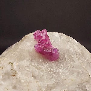 May include: A vibrant, raw ruby gemstone, displaying a deep pink hue, rests atop a white, textured mineral. The ruby's crystalline structure is visible, catching the light. The background is a dark, neutral tone, highlighting the gem's color and form.