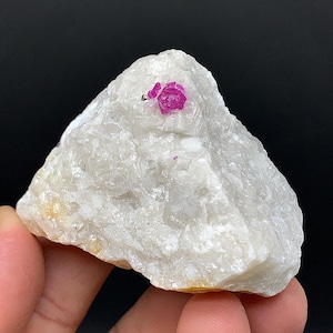 Peut inclure: Une pierre brute blanche avec un cristal de rubis rose vif incrusté dans sa surface. Le rubis est le point central, contrastant avec les tons neutres de la pierre. La pierre est tenue dans une main sur un fond noir.