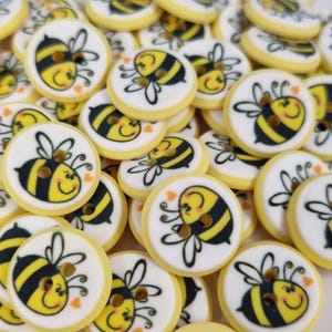 May include: A pile of circular buttons with a yellow edge and a white centre. Each button displays a cartoon bee with black and yellow stripes, a smiling face, and small heart details. The buttons have two holes for sewing.