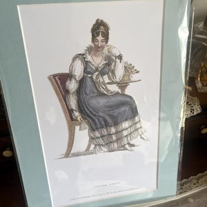 Peut inclure: Une impression encadrée d'une femme en robe bleue assise sur une chaise, avec une petite table à côté d'elle. L'impression est dans un cadre et un passe-partout bleu clair. Le texte "Dinner Paris" est en bas.