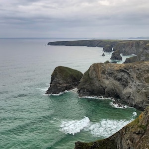 Puede incluir: Un paisaje costero escénico con una costa escarpada con acantilados y formaciones rocosas. El océano turquesa se encuentra con la orilla, con olas rompiendo contra las rocas. El cielo está nublado, creando una atmósfera melancólica.