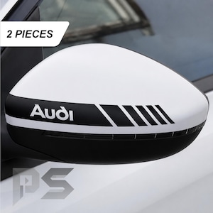 May include: A white and black car side mirror cover featuring the text Audi and a series of diagonal stripes. The set includes 2 pieces. The design consists of a white upper section and a black lower section with a graphic decal applied to the mirror housing.
