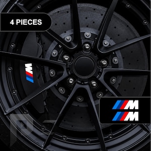 Op de afbeelding: Een set van 4 stuks M-logo stickers met het iconische blauwe, rode en witte kleurenschema. De stickers zijn ontworpen voor autoremklauwen en worden hier getoond op een zwart meerspaaks wiel met een donkere, geperforeerde remschijf.