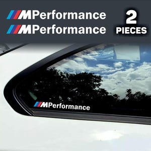 May include: White car window with a black trim, featuring a sticker with the text "MPerformance" in white, with blue and red stripes. The image also shows a second sticker with the same design and the text "2 PIECES".
