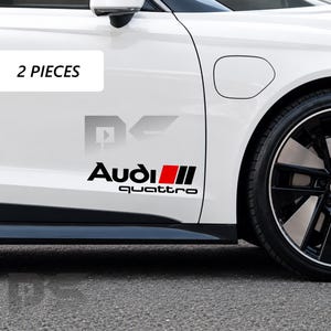 Può includere: Una portiera di auto bianca presenta un adesivo in vinile nero e rosso con il testo Audi quattro e una grafica a strisce rosse e nere. Un'etichetta rettangolare bianca nell'angolo in alto a sinistra mostra il testo nero 2 PIECES.