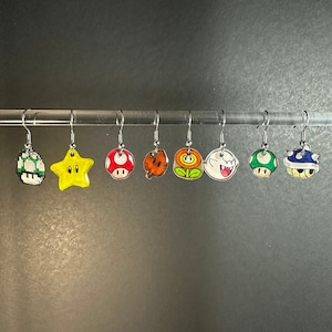 May include: Eight pairs of earrings featuring different characters from the Super Mario Bros. video game franchise. The earrings are made of metal and have a clear plastic charm with a painted design. The characters include a star, a mushroom, a flower, a ghost, a turtle shell, a koopa troopa, and a goomba.