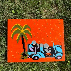 Puede incluir: Pintura de arte pixelado de una palmera y un vehículo azul claro sobre un fondo naranja. La obra de arte está compuesta por píxeles cuadrados, lo que le da una estética de videojuego retro. La pintura se muestra en un panel de madera.