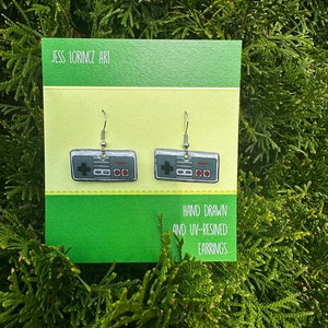 Puede incluir: Un par de pendientes de plata con un diseño de mando de videojuego clásico dibujado a mano y en resina UV. Los pendientes se muestran en una tarjeta verde y amarilla con el texto "JESS LORINCZ ART" y "HAND DRAWN AND UV-RESINED EARRINGS".