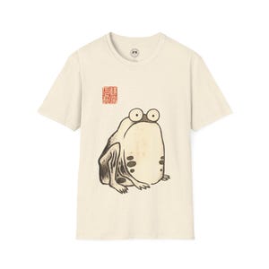 May include: Cream-colored t-shirt featuring a whimsical illustration of a frog with large eyes and a minimalist design. The frog is in shades of brown and beige. Above the frog is a red Japanese character stamp.