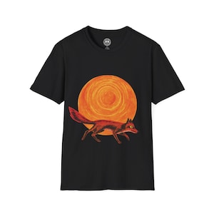 Puede incluir: Camiseta negra con una ilustración de un zorro rojo corriendo frente a un gran sol naranja. El zorro está representado en movimiento con una larga cola. El sol tiene un diseño circular texturizado. La camiseta tiene cuello redondo y mangas cortas.