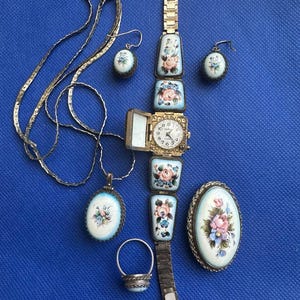 Puede incluir: Una colección de joyas vintage, que incluye un reloj, un collar, pendientes, un anillo y un broche. Cada pieza presenta un diseño floral con acentos rosas, azules y blancos sobre un fondo azul claro. El reloj tiene una correa de metal dorado.