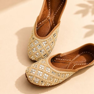May include: A pair of beige slip-on flats featuring a lattice pattern of small square mirrors and cream-colored beads. The interior is lined with brown material, and the edges are finished with a tan trim and visible white stitching detail.