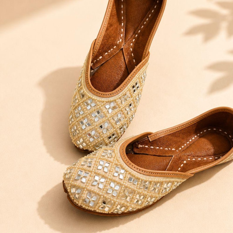 May include: A pair of beige slip-on flats featuring a lattice pattern of small square mirrors and cream-colored beads. The interior is lined with brown material, and the edges are finished with a tan trim and visible white stitching detail.