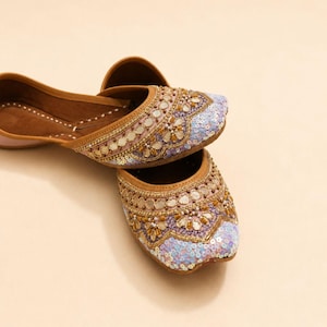 May include: A pair of flat slip-on shoes featuring a brown interior lining and an exterior adorned with intricate gold-toned embroidery, small reflective mirror accents, and a dense pattern of light blue and white sequins across the rounded toe.