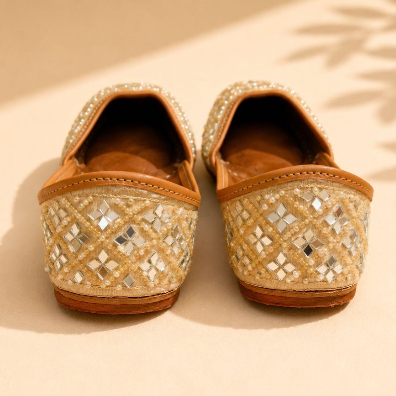 May include: A pair of beige slip-on flats featuring a diamond-patterned exterior adorned with small square mirrors and tiny white beads. The shoes have a tan leather-like trim along the opening and a solid brown sole, showcasing a detailed texture.