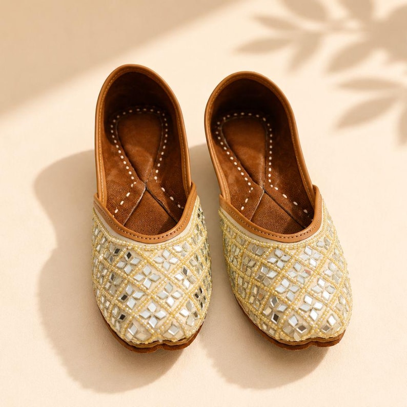 May include: A pair of slip-on flats featuring a cream-colored upper adorned with a diamond-shaped grid of small mirrors and white beads. The interior is lined with brown material, and the edges are finished with a tan trim around the opening.
