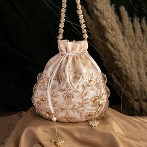 Puede incluir: Un bolso con cordón de color crema con intrincados bordados florales dorados. El bolso presenta un asa con cuentas de perlas y borlas. El diseño incluye un borde festoneado y una textura similar a la seda, creando un accesorio elegante.