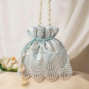 Könnte beinhalten: Hellblaue Satin-Kordelzugtasche mit aufwendiger Perlen- und Silberperlenarbeit. Die Tasche hat einen gewellten Saum und einen Perlenhenkel. Eine goldene Quaste mit Perlen hängt an der Seite. Ein stilvolles Accessoire.