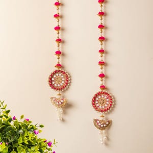 May include: Two decorative hanging ornaments. Each ornament features a circular design with a red and gold border, a white center, and a gold pendant with white beads. The ornaments are adorned with pink and gold beads.