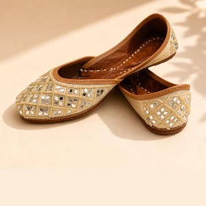 May include: A pair of beige flat shoes featuring a diamond-patterned exterior adorned with small square mirrors and delicate beadwork. The shoes have a brown leather interior lining and a rounded toe design, showcasing intricate craftsmanship.