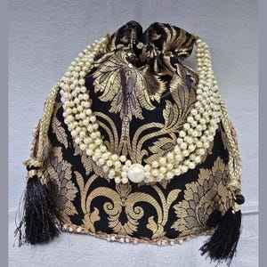 May include: A black and gold patterned drawstring bag with a pearl necklace design. The bag features a floral pattern, black tassels, and a large pearl accent. The drawstring is gold, and the bag is embellished with small pearls.