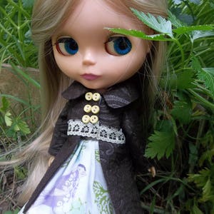 May include: A doll with long blonde hair and large blue eyes, dressed in a dark brown coat with yellow buttons and white lace trim. The doll's dress has a bird and leaf pattern. Green foliage forms the background.