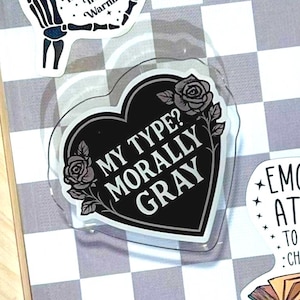May include: Three vinyl stickers on a gray and white checkered surface. One sticker shows skeleton hands forming a heart with the text You had me at Trigger Warnings. Another is a black heart with roses and the text My Type? Morally Gray. The third is an open book with the text Emotionally Attached To Fictional Characters.