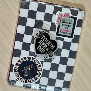 May include: A clear protective case for an e-reader with a black and white checkered pattern. The case is decorated with stickers, including a heart-shaped sticker with the text "TOUCH HER & DIE" and a sticker that says "if I die please delete my kindle history".