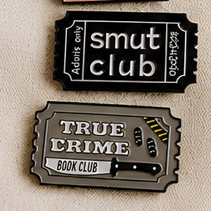 May include: Two rectangular enamel pins shaped like admission tickets. The top pin is black with white text reading 'smut club' and 'Adonis only'. The bottom pin is gray with white text reading 'TRUE CRIME BOOK CLUB' and a graphic of a kitchen knife.