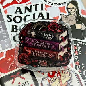 May include: A clear acrylic sticker featuring a stack of three books with dark covers. The titles read The Lady & the Orc, Guarded by the Gargoyle, and Claimed by the Dragon. The design includes red roses, a clawed hand, and a heart-shaped gem.