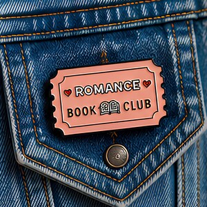 May include: A rectangular enamel pin shaped like a ticket with a light pink background, black text reading ROMANCE BOOK CLUB, and two small red hearts. The pin is attached to a blue denim fabric pocket with orange stitching and a single metal button.