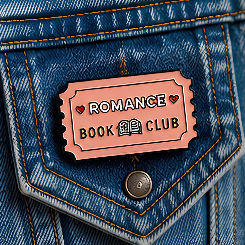 May include: A rectangular enamel pin shaped like a ticket with a light pink background, black text reading ROMANCE BOOK CLUB, and two small red hearts. The pin is attached to a blue denim fabric pocket with orange stitching and a single metal button.