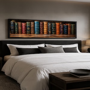 May include: A rectangular wall art piece featuring a row of antique books with colourful spines in shades of blue, red, green, and orange. The artwork is displayed in a black frame above a bed with white bedding, dark pillows, and two bedside lamps.