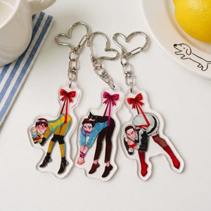 May include: Three clear acrylic keychains featuring illustrated characters suspended by red ribbons. Each keychain has a heart-shaped metal clasp. The characters are depicted in various outfits, including a yellow jumpsuit, a blue shirt, and a maid uniform.