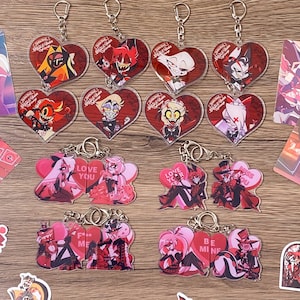 May include: A collection of heart-shaped acrylic keychains featuring characters from Hazbin Hotel. The top row displays individual character portraits on red backgrounds, while the bottom rows show pairs with text including Love You, Miss You, F*** Me, and Be Mine.