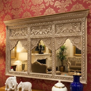 May include: An intricately carved, off-white wooden mirror with three panels, reflecting a room with furniture and a fireplace. Two white elephant ornaments and a blue vase are on a wooden surface. The mirror is mounted on a red patterned wall.
