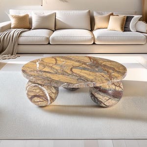 May include: A circular coffee table with a marble top and three spherical legs. The table's surface has brown, tan, and grey veining. The legs are also marble, with similar colour variations. The table rests on a white rug.