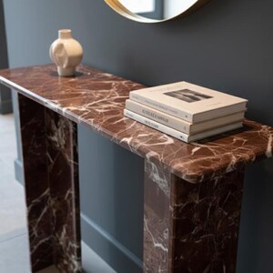 Puede incluir: Una consola rectangular con tapa de m&aacute;rmol y dos patas de soporte. La mesa es de color marr&oacute;n rojizo oscuro con vetas blancas. Una pila de libros y un jarr&oacute;n blanco est&aacute;n sobre la mesa. Un espejo con marco dorado cuelga de la pared.