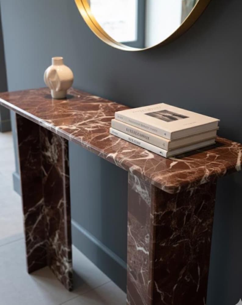 Puede incluir: Una consola rectangular con tapa de m&aacute;rmol y dos patas de soporte. La mesa es de color marr&oacute;n rojizo oscuro con vetas blancas. Una pila de libros y un jarr&oacute;n blanco est&aacute;n sobre la mesa. Un espejo con marco dorado cuelga de la pared.