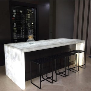 May include: A modern bar with a white, illuminated countertop and black metal stools. The countertop has a marble-like appearance. A wine rack is visible in the background, along with a decorative glass. The stools have a woven seat design.