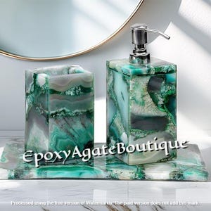 Puede incluir: Un juego de baño con un dispensador de jabón, un vaso y una bandeja de piedra de ágata verde y gris. El dispensador de jabón tiene una bomba plateada. El juego se exhibe sobre una superficie blanca.