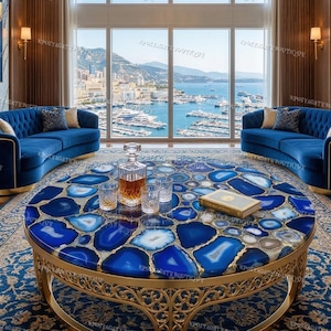 Puede incluir: Un salón lujoso con una mesa de centro redonda con una tapa de piedra de ágata azul y una base de metal dorado. Dos sofás curvos de terciopelo azul están a ambos lados de la mesa. La habitación tiene una gran ventana con vistas a un puerto.