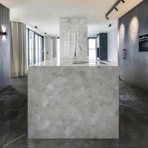 May include: A modern kitchen island with a light gray, patterned countertop and matching base. The island features a built-in sink with a curved faucet. The kitchen has dark gray cabinets, a black floor, and large windows.
