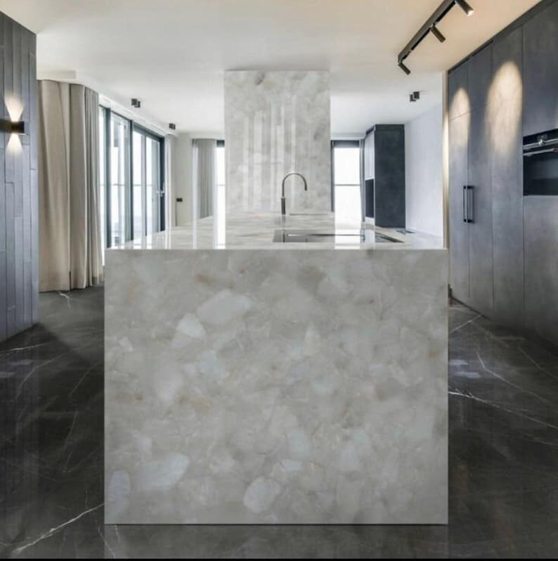 May include: A modern kitchen island with a light gray, patterned countertop and matching base. The island features a built-in sink with a curved faucet. The kitchen has dark gray cabinets, a black floor, and large windows.