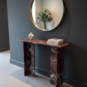 Puede incluir: Una consola de mármol rojo con un espejo redondo con marco dorado encima. La mesa tiene una tapa rectangular y dos patas rectangulares. Un pequeño jarrón blanco y libros están sobre la mesa. El espejo refleja una planta en maceta. La mesa mide unos 120 cm de largo.