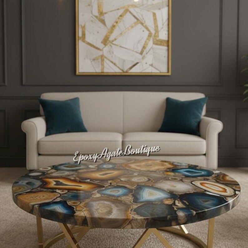 May include: A round coffee table with a unique agate stone top in shades of blue, brown, and gold. The table has a gold metal base and is set in a living room with a cream-colored sofa and teal pillows. A framed abstract artwork hangs on the wall.