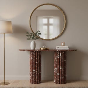 May include: A modern console table with a red marble top and cylindrical legs. A gold-framed round mirror hangs above the table. A white vase with greenery, books, and a candle are on the table. A gold floor lamp is to the left.